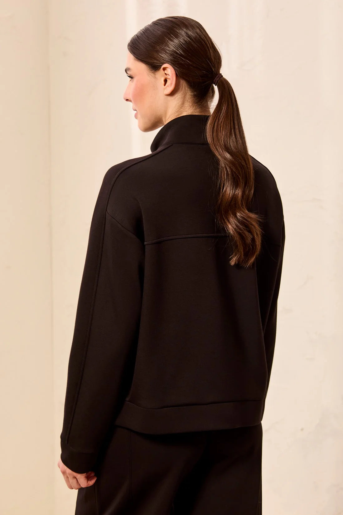 A woman shown from the back wearing a black cropped jacket with a mock neck and long sleeves, paired with black bottoms, against a light beige textured wall.