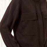 A close-up of a person wearing a dark brown or very deep espresso zip-front jacket with a stand collar, long sleeves, and two large flap chest pockets.