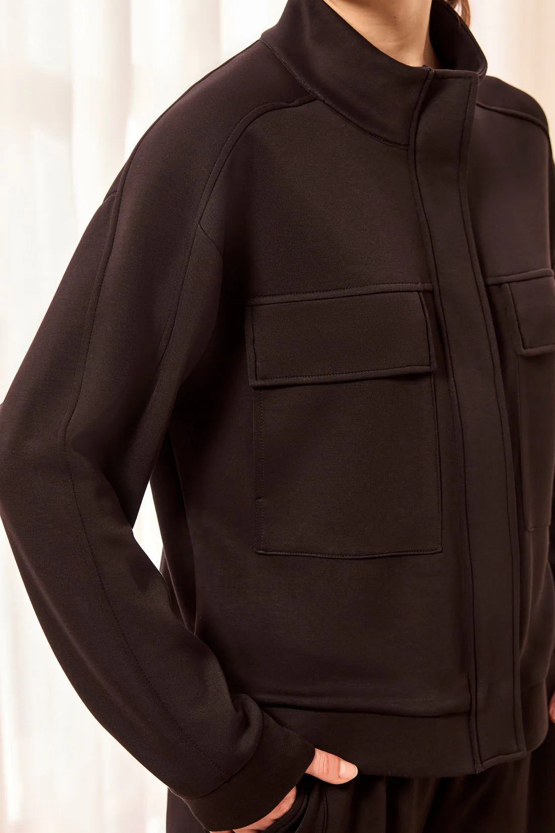 A close-up of a person wearing a dark brown or very deep espresso zip-front jacket with a stand collar, long sleeves, and two large flap chest pockets.