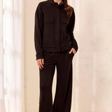 A woman modeling a black long-sleeve zip-up jacket with flap chest pockets and matching black wide-leg pants, standing in a bright indoor setting with white curtains and a light wood floor.