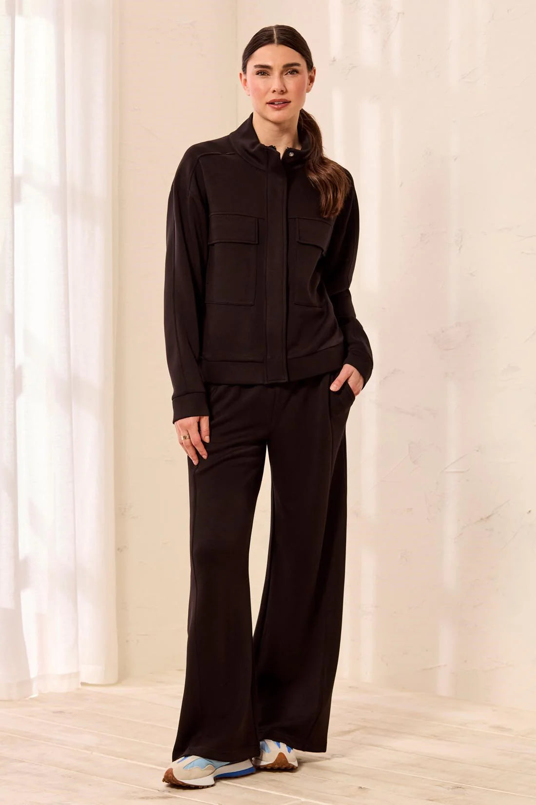 A woman modeling a black long-sleeve zip-up jacket with flap chest pockets and matching black wide-leg pants, standing in a bright indoor setting with white curtains and a light wood floor.