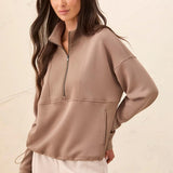 A woman modeling a taupe brown quarter-zip pullover sweatshirt with a mock neck and zippered hand pockets, paired with cream pants against a textured beige wall.
