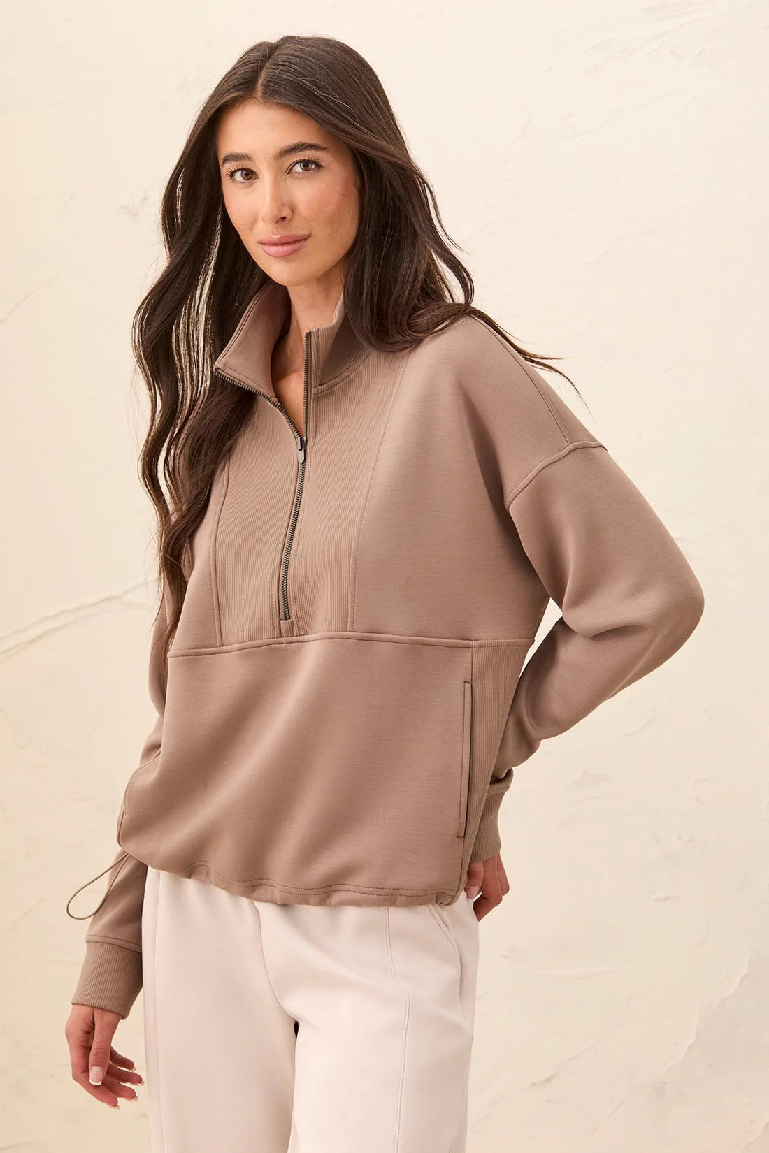 A woman modeling a taupe brown quarter-zip pullover sweatshirt with a mock neck and zippered hand pockets, paired with cream pants against a textured beige wall.
