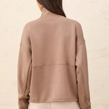 A woman shown from the back wearing a taupe-brown mock neck long-sleeve top and light beige pants, standing against a light textured wall.