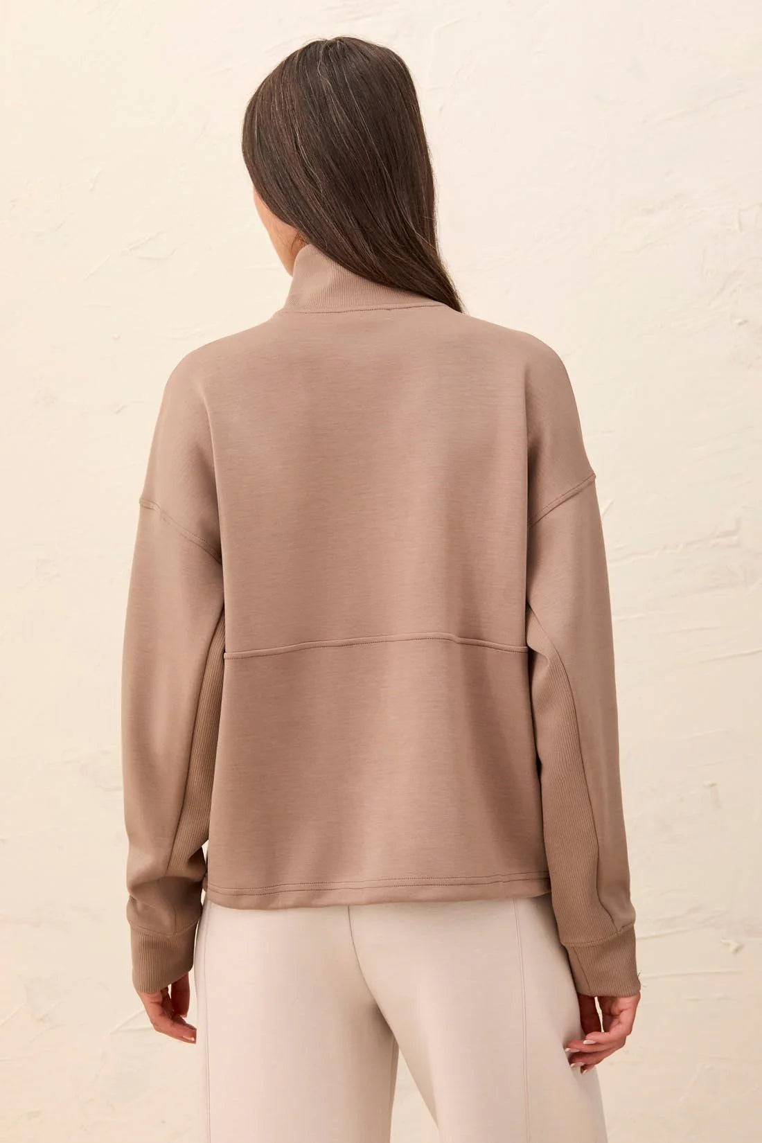 A woman shown from the back wearing a taupe-brown mock neck long-sleeve top and light beige pants, standing against a light textured wall.