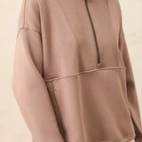 A close-up of a person wearing a taupe-beige quarter-zip pullover sweatshirt with a mock neck and front pockets, paired with light beige pants against a neutral cream background.