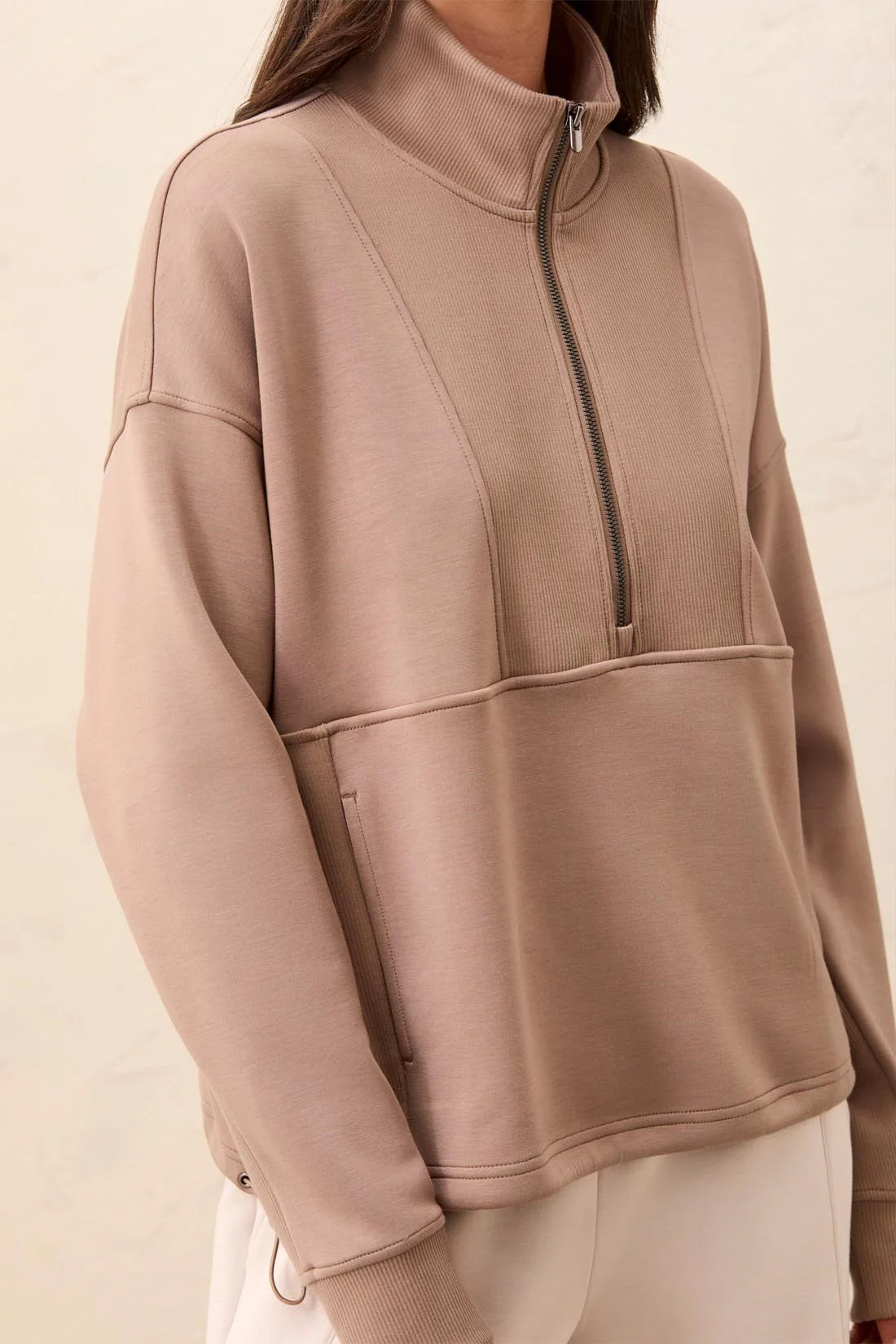 A close-up of a person wearing a taupe-beige quarter-zip pullover sweatshirt with a mock neck and front pockets, paired with light beige pants against a neutral cream background.