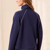 A woman shown from the back wearing a navy blue mock-neck athletic jacket with thin white piping details and matching navy pants, set against a neutral cream-colored background.