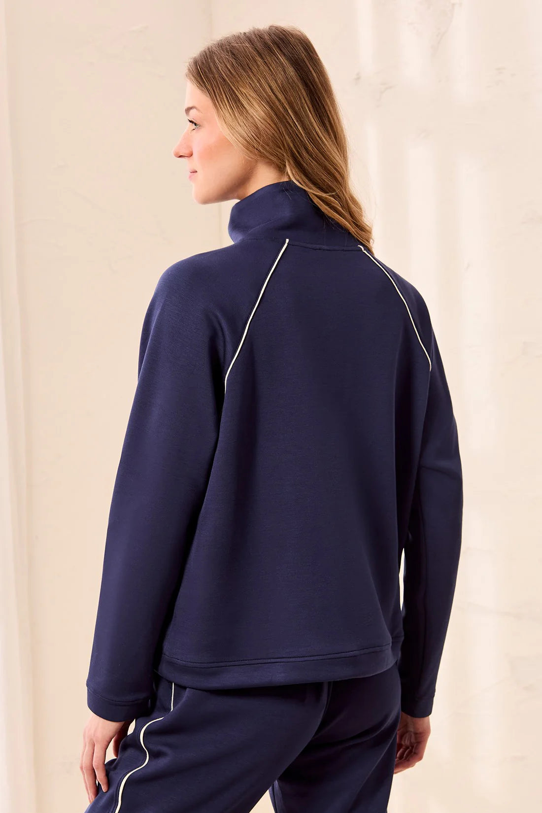 A woman shown from the back wearing a navy blue mock-neck athletic jacket with thin white piping details and matching navy pants, set against a neutral cream-colored background.
