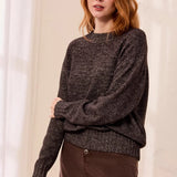A woman with red hair wearing a dark brown textured knit sweater and a brown skirt, standing against a soft cream-colored curtain background.