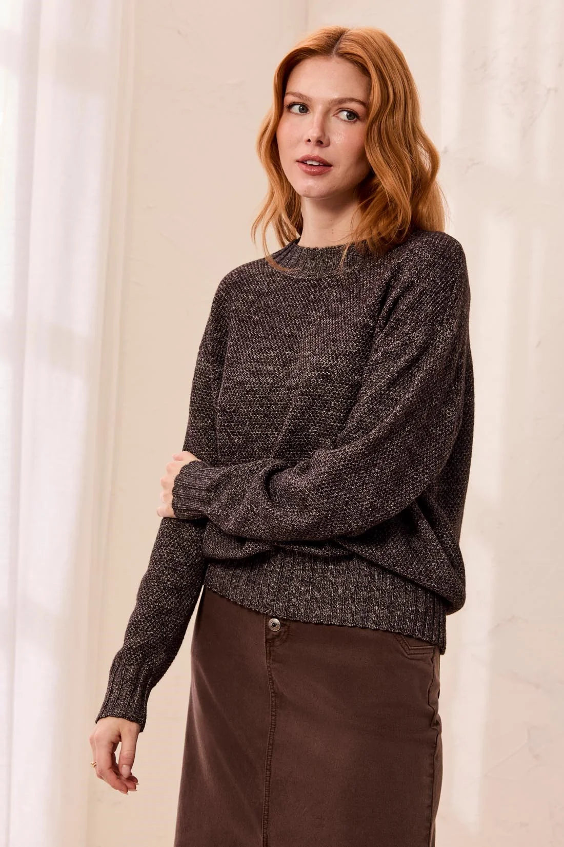 A woman with red hair wearing a dark brown textured knit sweater and a brown skirt, standing against a soft cream-colored curtain background.