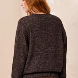 A woman with red hair is shown from the back wearing a dark brown textured knit sweater and brown pants against a light beige wall.