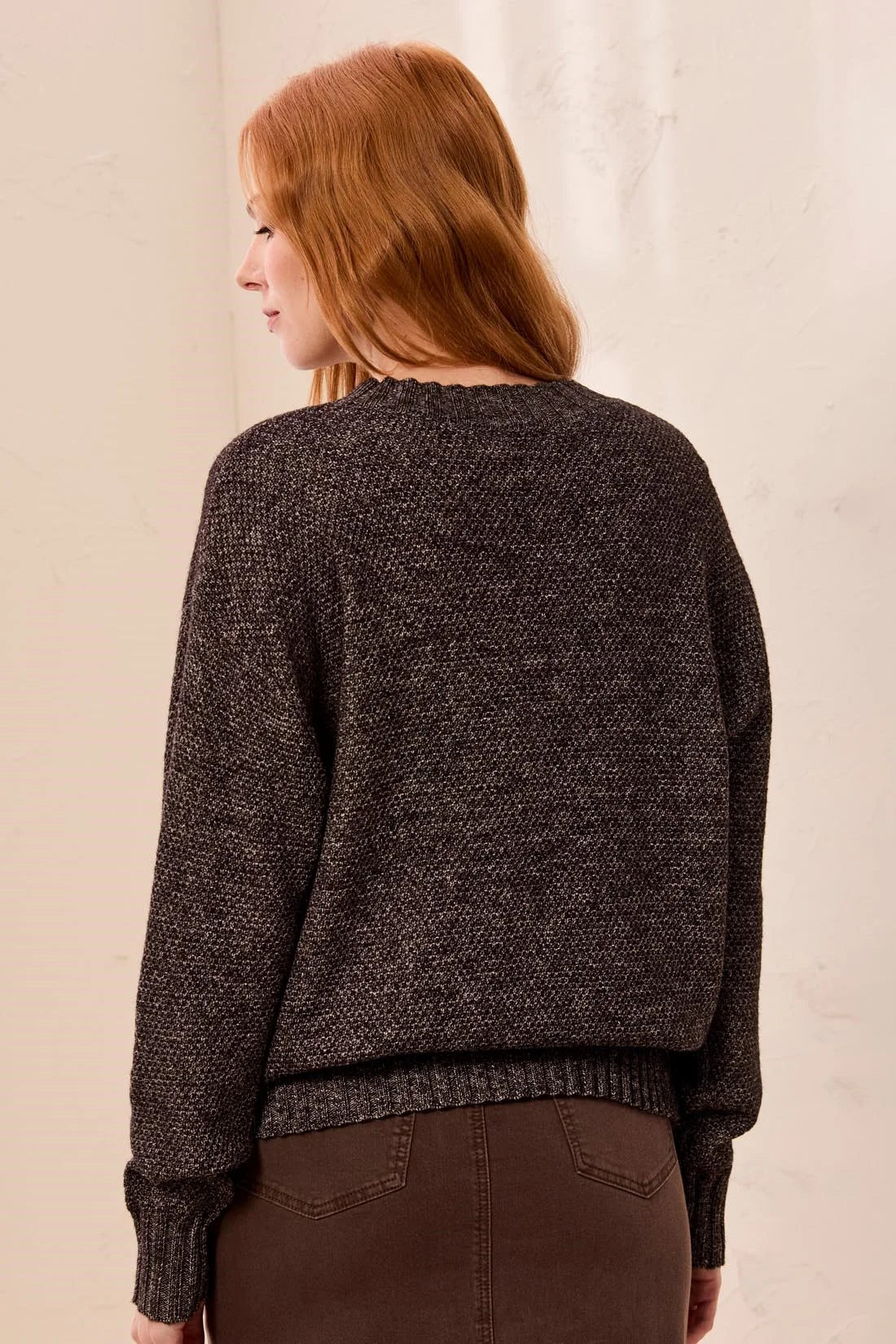 A woman with red hair is shown from the back wearing a dark brown textured knit sweater and brown pants against a light beige wall.
