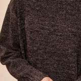 A close-up of a person wearing a dark brown and black heathered knit crewneck sweater, with hands clasped in front against a plain light beige background.