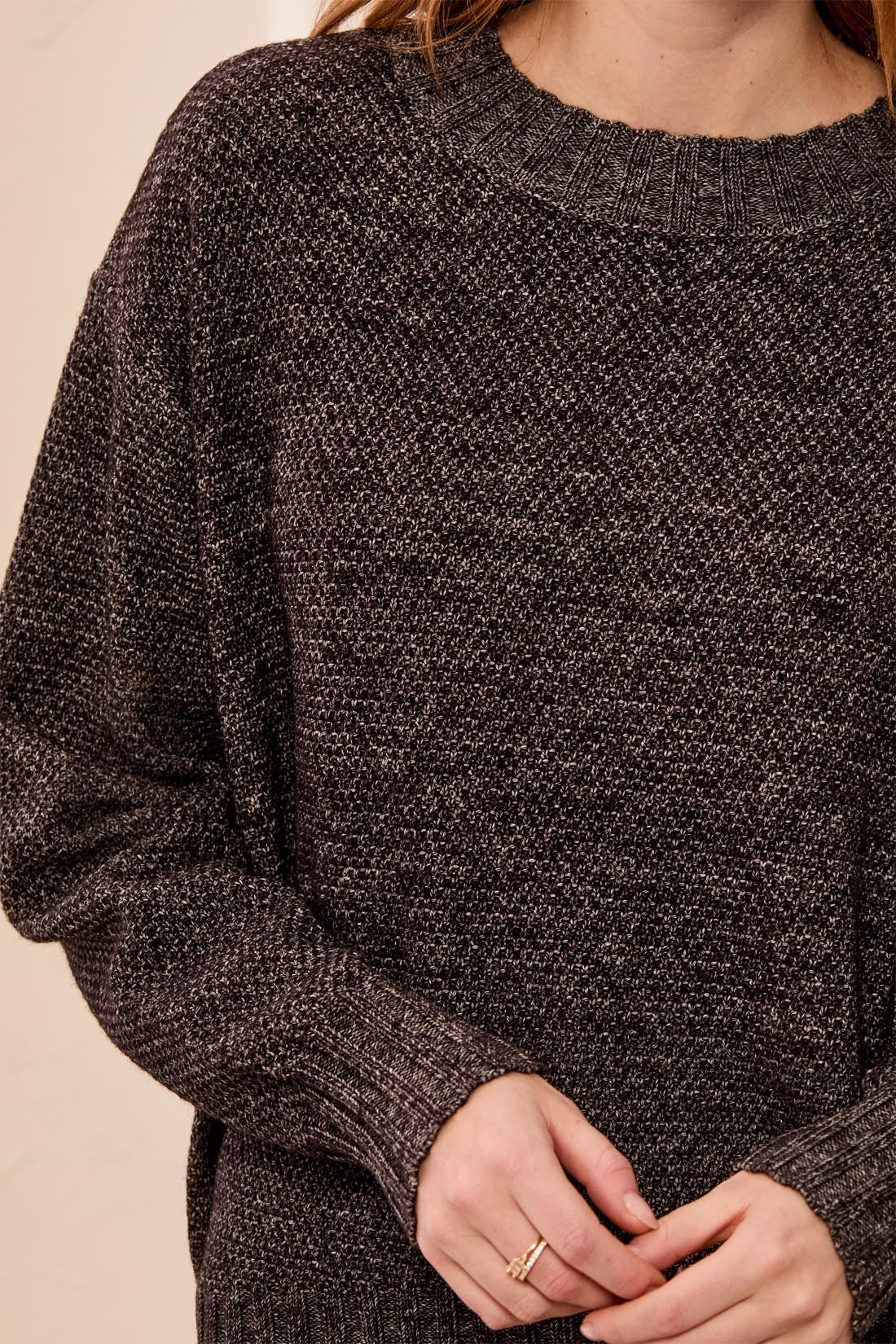 A close-up of a person wearing a dark brown and black heathered knit crewneck sweater, with hands clasped in front against a plain light beige background.