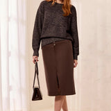 A woman stands in a bright indoor setting wearing a dark charcoal gray knit sweater and a brown knee-length skirt, holding a small dark handbag and wearing brown pointed flats.