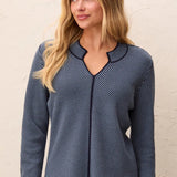 A woman wearing a navy blue long-sleeve top with thin white diagonal stripes and a scalloped V-neckline, standing against a textured off-white wall.