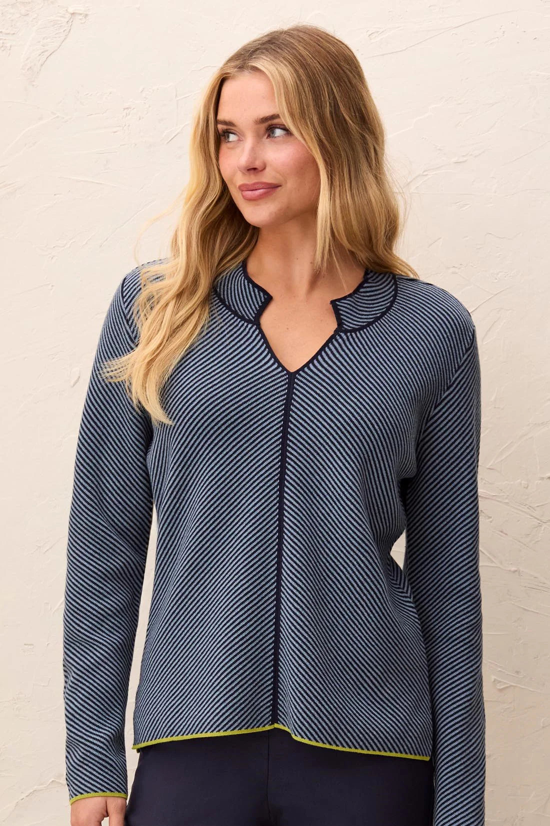 A woman wearing a navy blue long-sleeve top with thin white diagonal stripes and a scalloped V-neckline, standing against a textured off-white wall.
