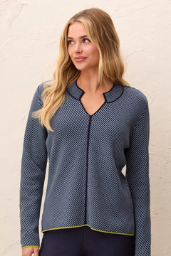 A woman wearing a navy blue long-sleeve top with thin white diagonal stripes and a scalloped V-neckline, standing against a textured off-white wall.