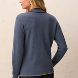 A woman shown from the back wearing a navy blue and white striped long-sleeve knit top with a thin yellow-green hem trim, paired with dark pants, against a textured off-white wall.
