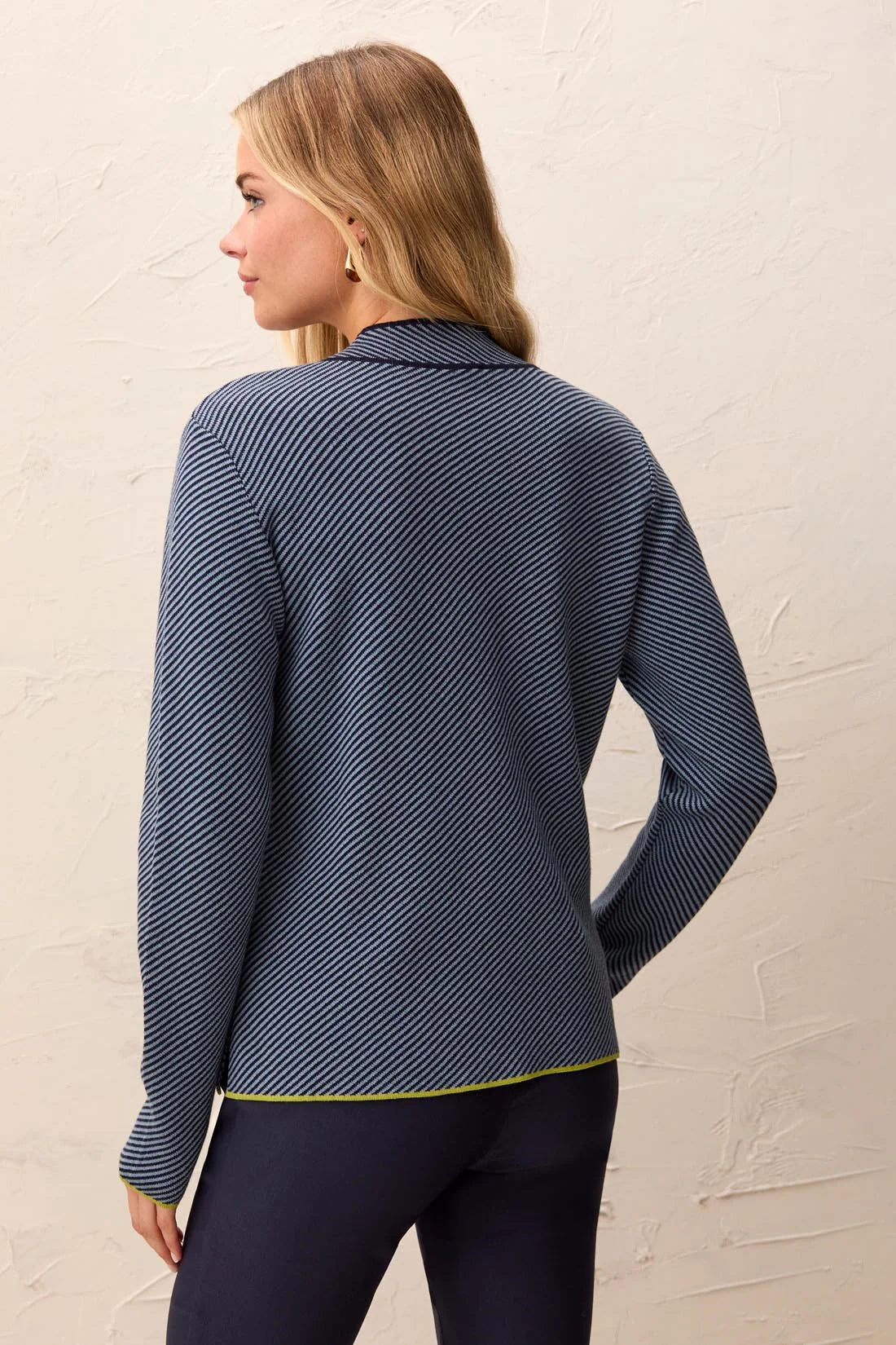 A woman shown from the back wearing a navy blue and white striped long-sleeve knit top with a thin yellow-green hem trim, paired with dark pants, against a textured off-white wall.
