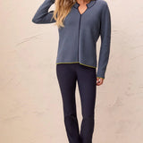 A woman modeling a two-piece loungewear set consisting of a slate blue long-sleeve knit top with a subtle pattern and dark navy pants, standing against a textured beige wall.