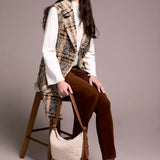 Woman sitting on a stool wearing a plaid vest, white shirt, brown pants, and brown boots, holding a white handbag.
