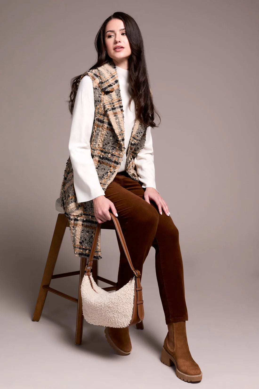 Woman sitting on a stool wearing a plaid vest, white shirt, brown pants, and brown boots, holding a white handbag.