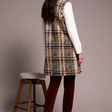 Woman wearing a plaid dress with a white shirt underneath, standing next to a wooden stool against a plain background.