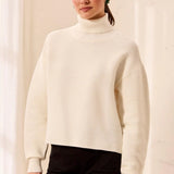A woman wearing a cream ribbed turtleneck sweater and black pants, standing against a soft beige curtain background.