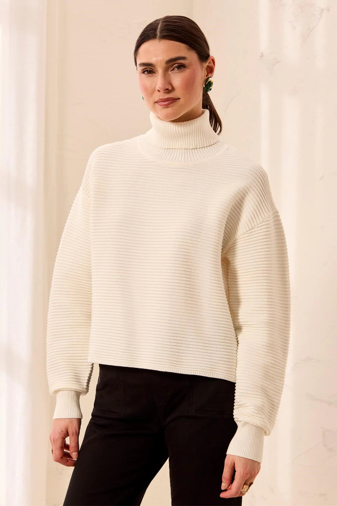 A woman wearing a cream ribbed turtleneck sweater and black pants, standing against a soft beige curtain background.