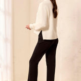 A woman stands in profile wearing a cream ribbed turtleneck sweater, black trousers, and black ankle boots with wooden block heels, posed in a bright indoor room with light-colored walls and floor.