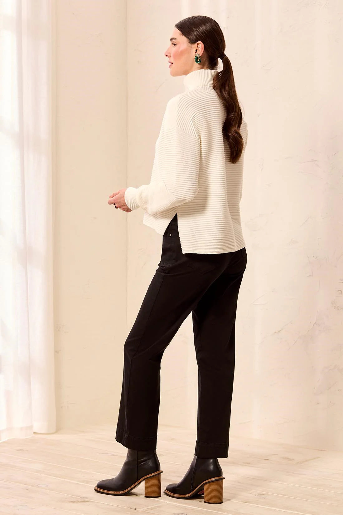 A woman stands in profile wearing a cream ribbed turtleneck sweater, black trousers, and black ankle boots with wooden block heels, posed in a bright indoor room with light-colored walls and floor.