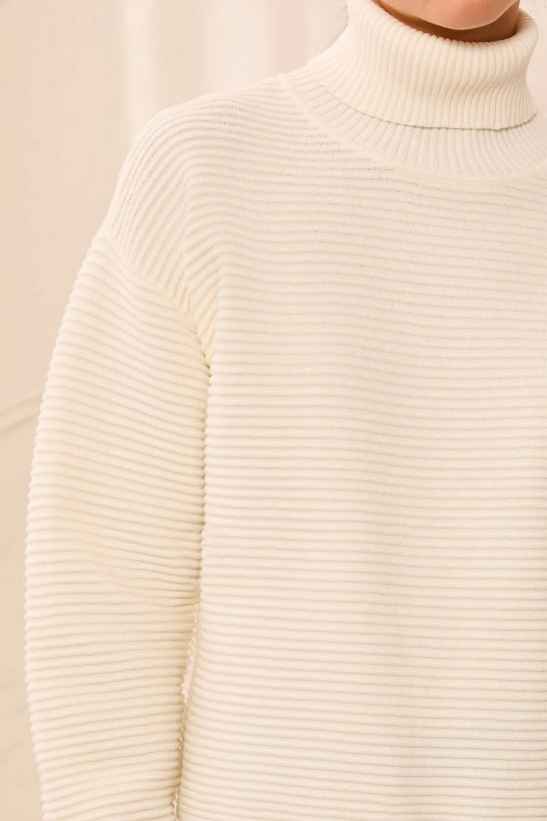 Close-up of a cream ribbed turtleneck sweater worn by a model, showing the fine horizontal ribbed texture of the knit fabric.