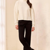 A woman modeling a ribbed cream turtleneck sweater and black trousers with black ankle boots, standing in a bright, minimalist room with a textured beige wall.
