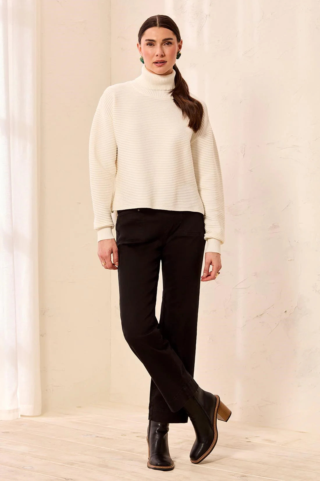A woman modeling a ribbed cream turtleneck sweater and black trousers with black ankle boots, standing in a bright, minimalist room with a textured beige wall.