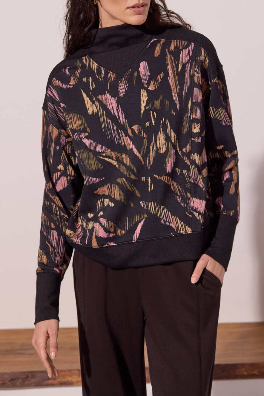 A woman wearing a black long-sleeve mock-neck sweatshirt with a pink and gold abstract brushstroke pattern, paired with black pants.