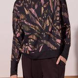 A woman wearing a black long-sleeve mock-neck sweatshirt with a pink and gold abstract brushstroke pattern, paired with black pants.
