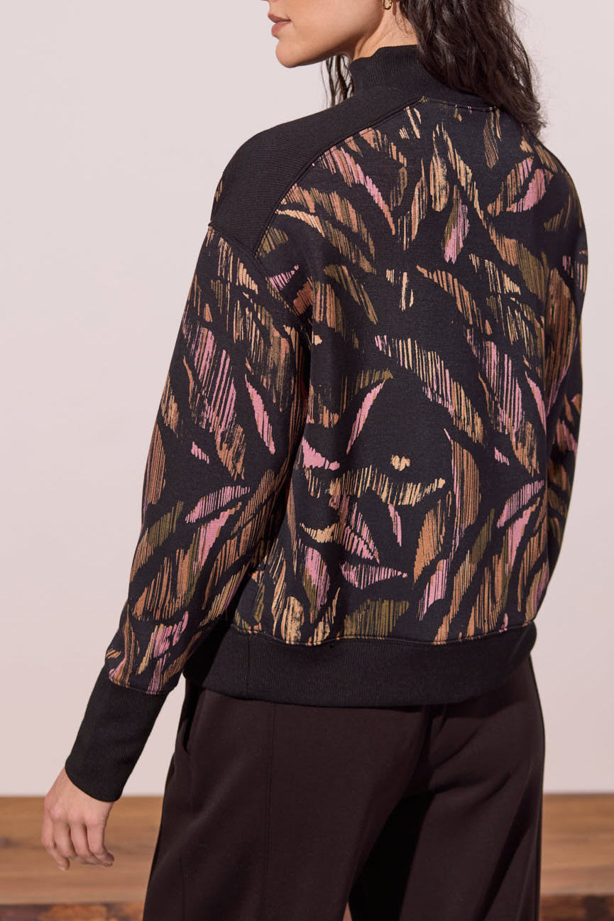 A woman shown from the back wearing a top with a gold and pink abstract leaf pattern, paired with black pants.