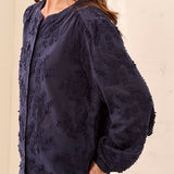A woman wearing a dark navy blue textured blouse with voluminous balloon sleeves, paired with light gray pants, shown against a plain beige background.