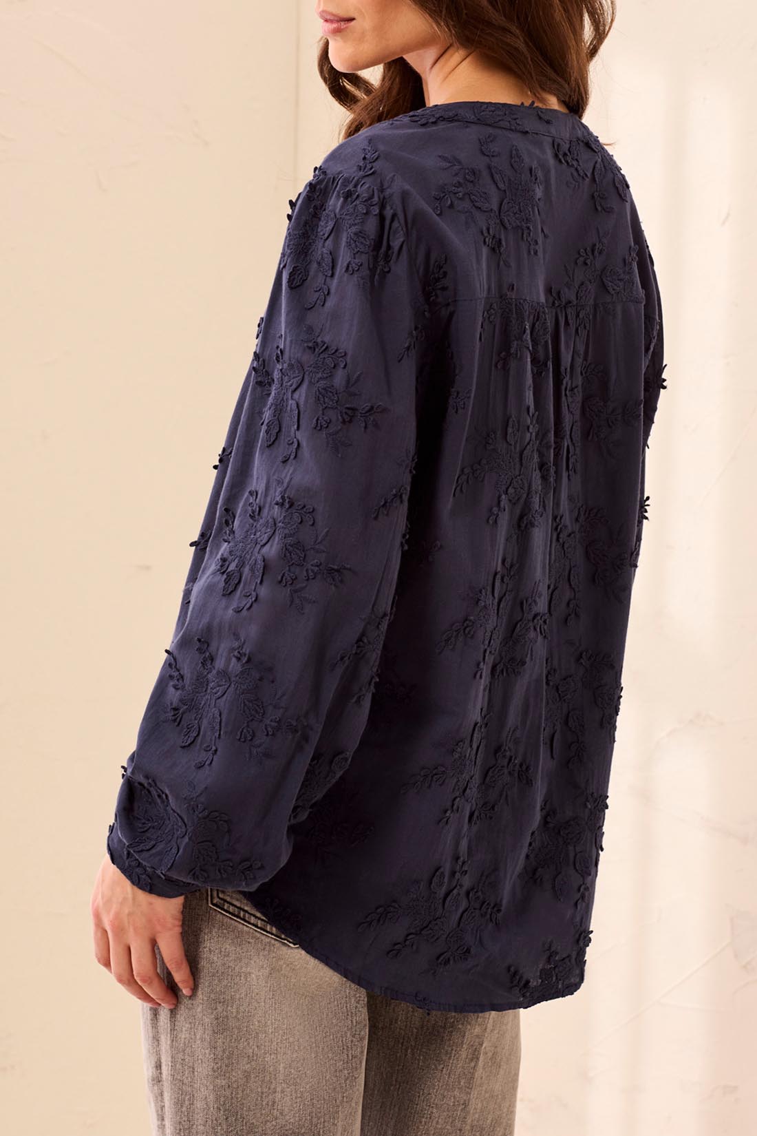 A woman wearing a dark navy blue long-sleeve blouse with intricate floral embroidery and textured applique, paired with light gray jeans, shown from a side-back angle against a neutral cream background.