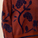 Woman wearing a  brownish orange sweater with floral blue detailing against a light back ground.