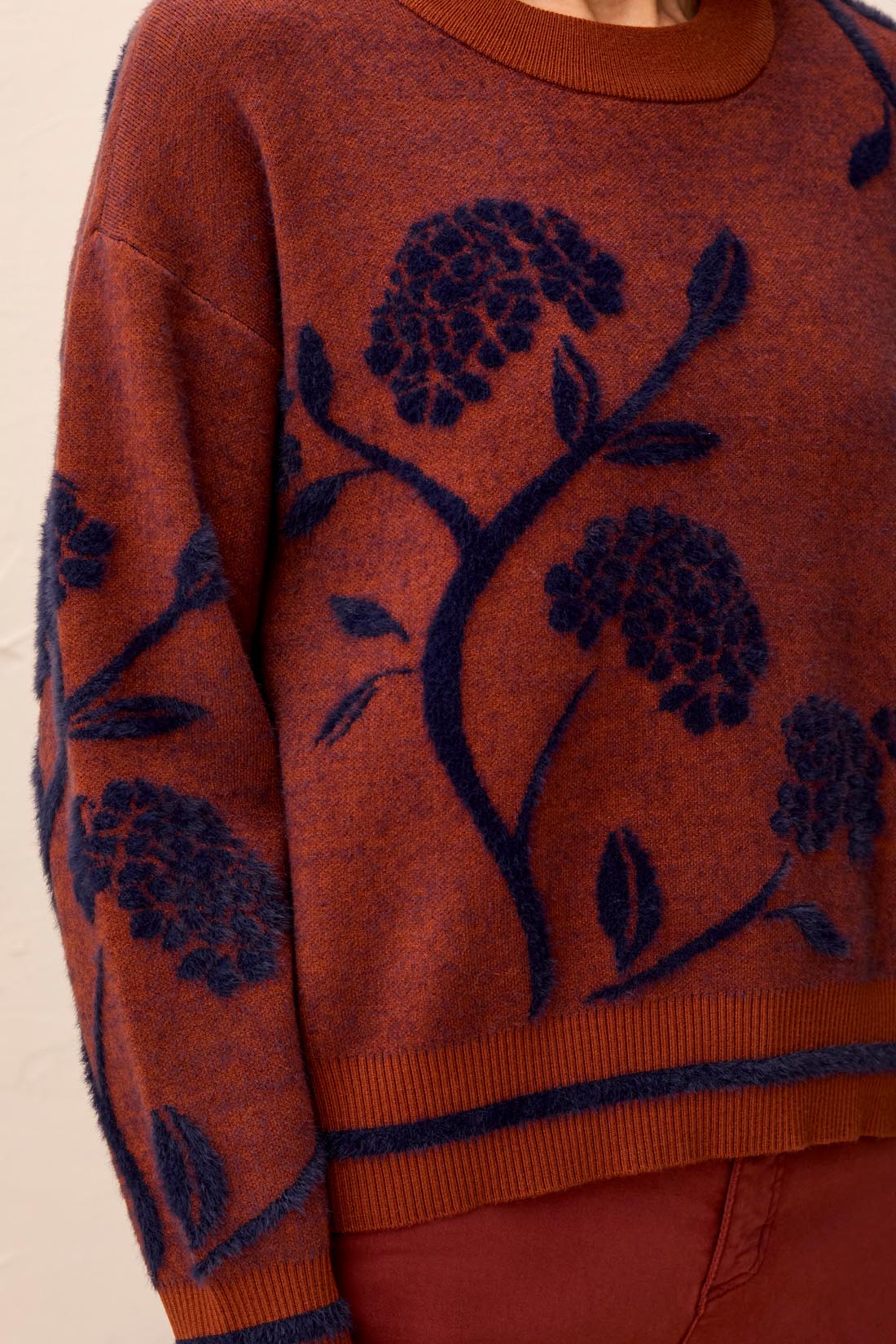 Woman wearing a  brownish orange sweater with floral blue detailing against a light back ground.