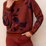 Woman wearing a  brownish orange sweater with floral blue detailing against a light back ground.