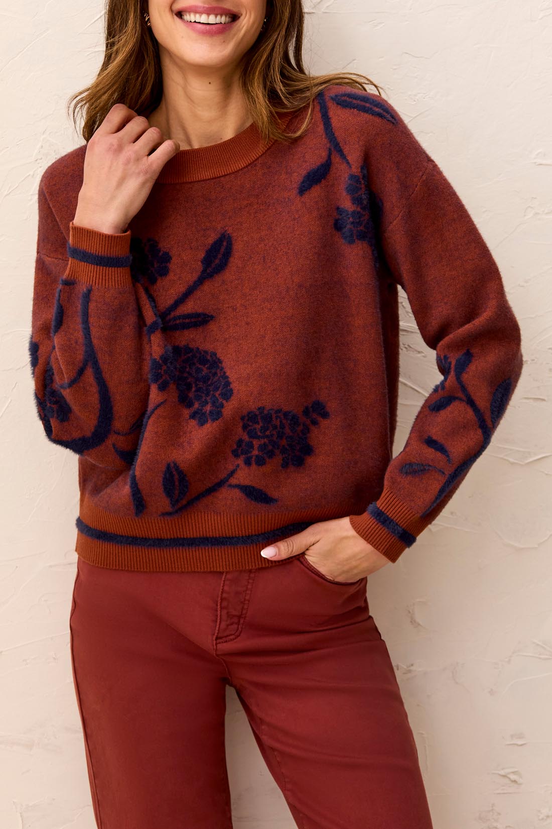 Woman wearing a  brownish orange sweater with floral blue detailing against a light back ground.