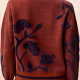 Woman wearing a  brownish orange sweater with floral blue detailing against a light back ground.