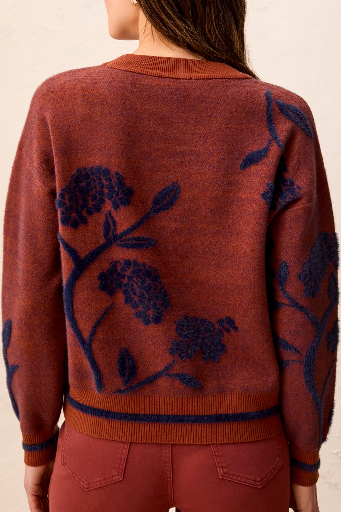 Woman wearing a  brownish orange sweater with floral blue detailing against a light back ground.