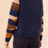 A woman standing with her back to the photographer, wearing a chunky knit cardigan sweater with a solid navy blue on the back and rust orange, cream, and dark blue on the sleeve.