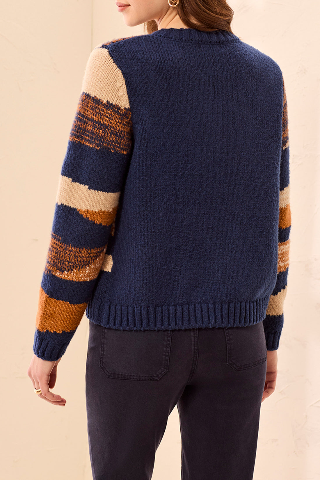 A woman standing with her back to the photographer, wearing a chunky knit cardigan sweater with a solid navy blue on the back and rust orange, cream, and dark blue on the sleeve.