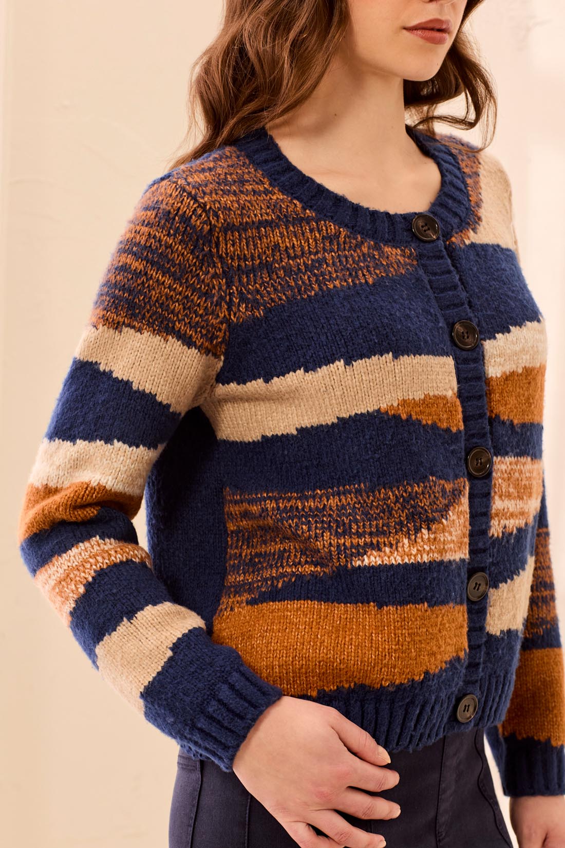 A woman wearing a chunky knit cardigan sweater with horizontal stripes in navy blue, rust orange, and cream, featuring dark brown buttons down the front.